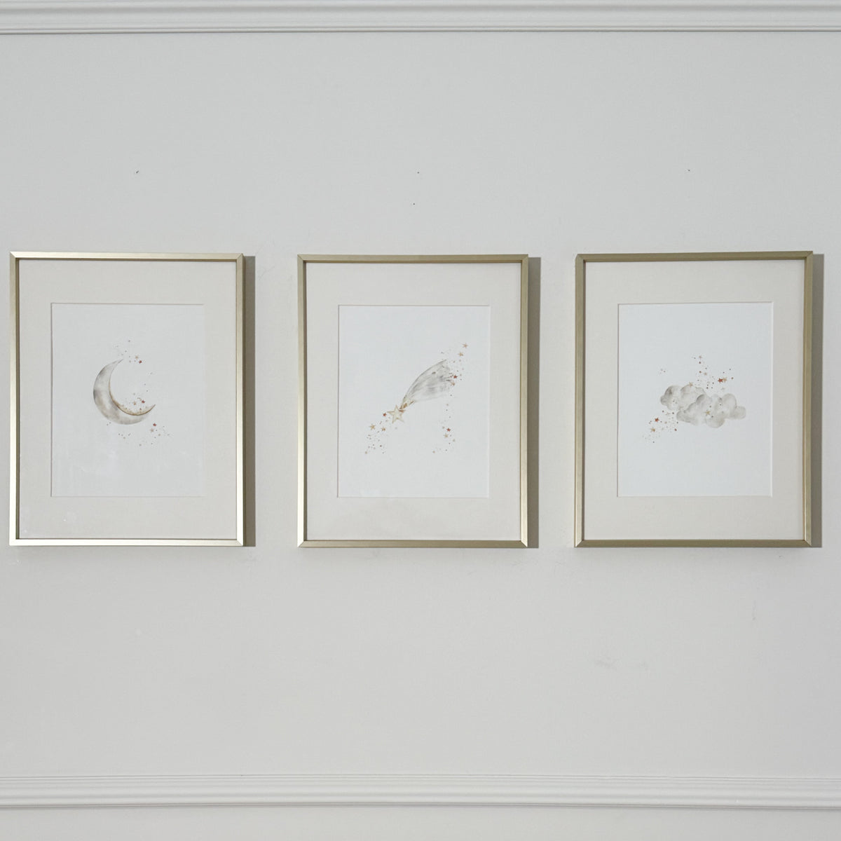 Three framed watercolor art prints with gold frames hanging side-by-side on a white wall. The prints feature minimalist designs of a crescent moon, a shooting star, and clouds.