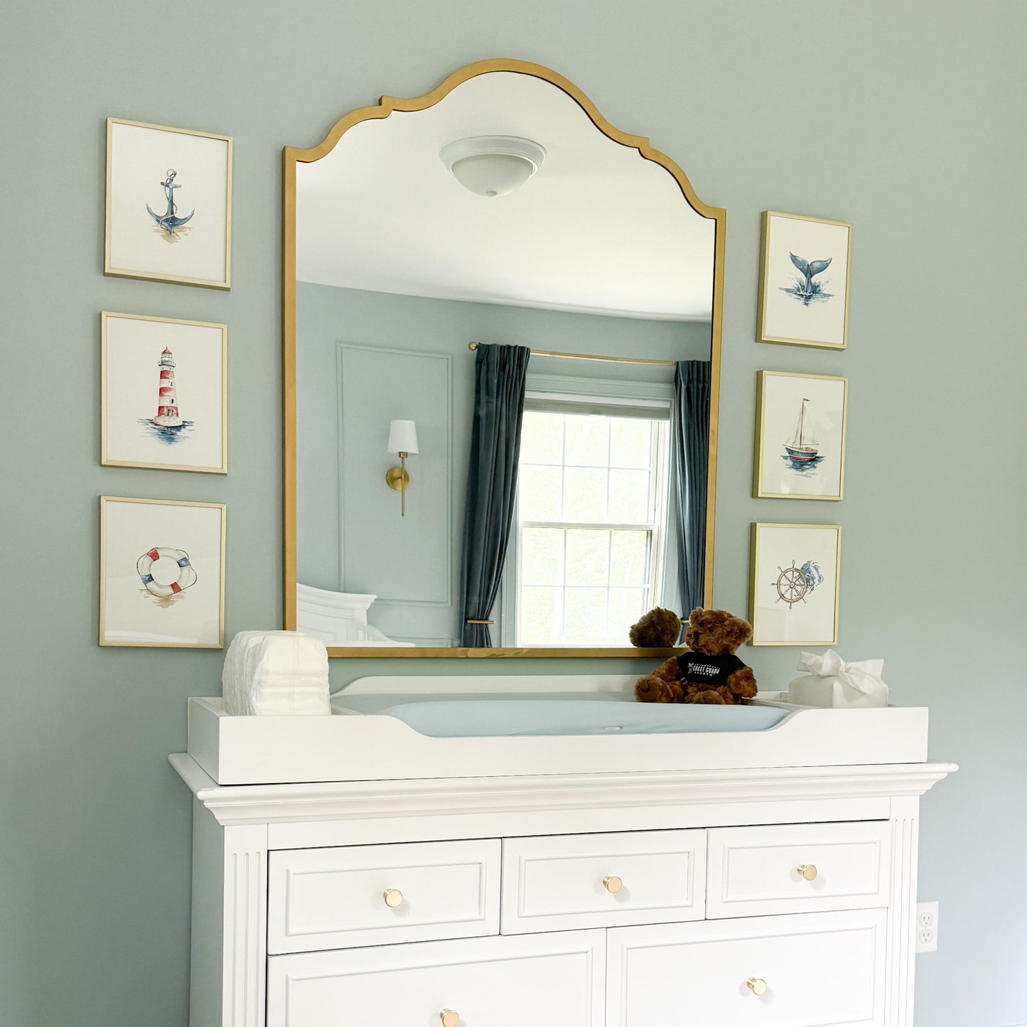 A large arched gold-framed mirror mounted on a pale blue wall above a white dresser, with framed nautical prints on both sides and a changing tray and teddy bear on the mirror surface.
