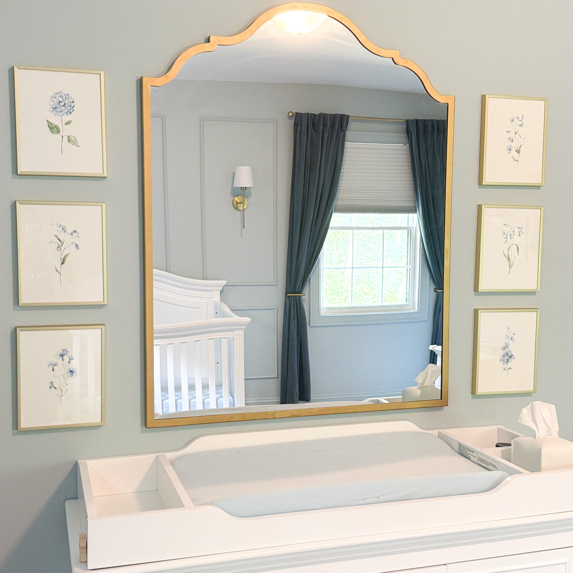 A large arched gold-framed nursery mirror mounted on a light blue-gray wall, flanked by six framed botanical prints. The mirror reflects a white changing crib, a window with dark teal curtains, and a gold wall sconce.