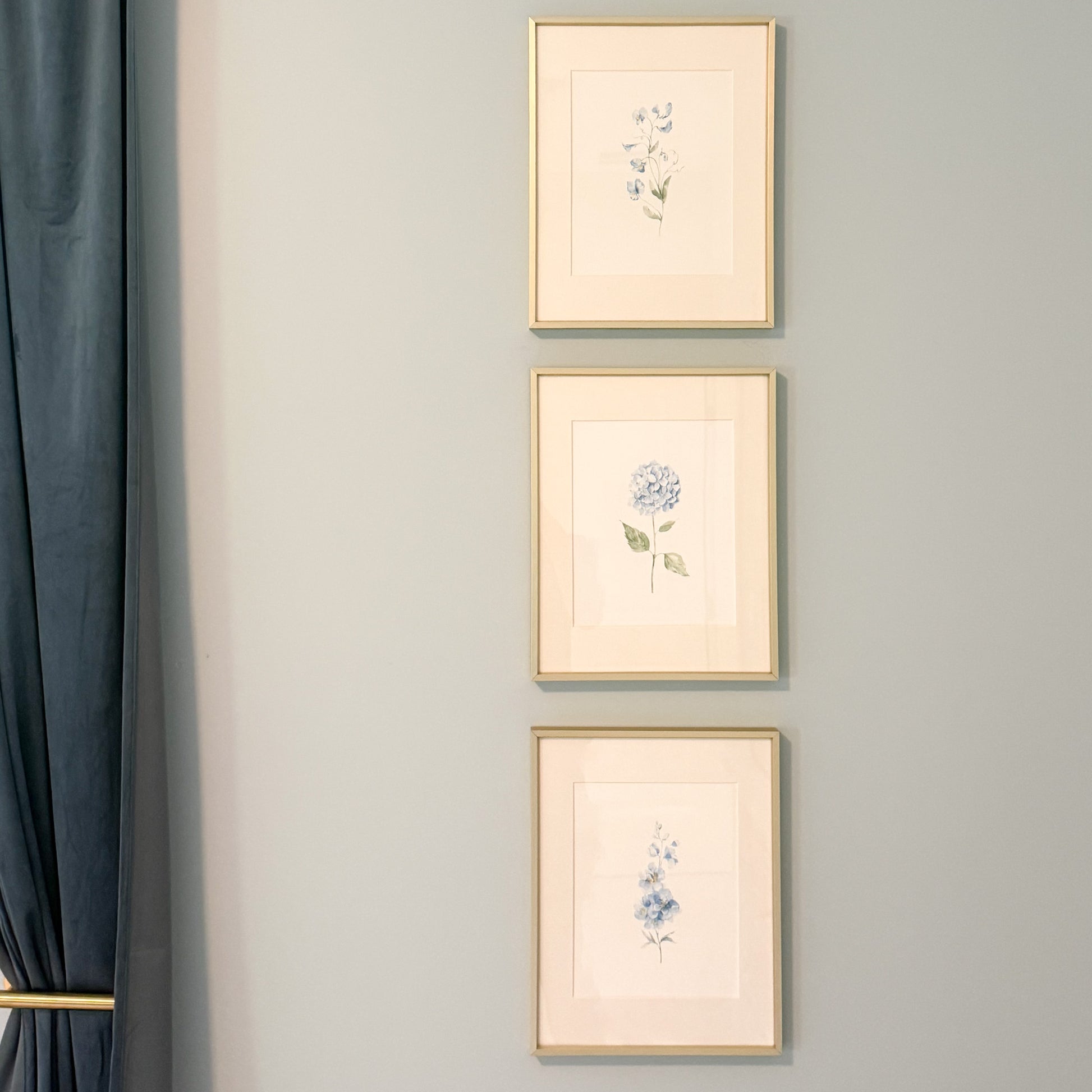 Three vertically aligned framed blue botanical art prints with gold frames hang on a light blue wall next to a dark blue curtain.