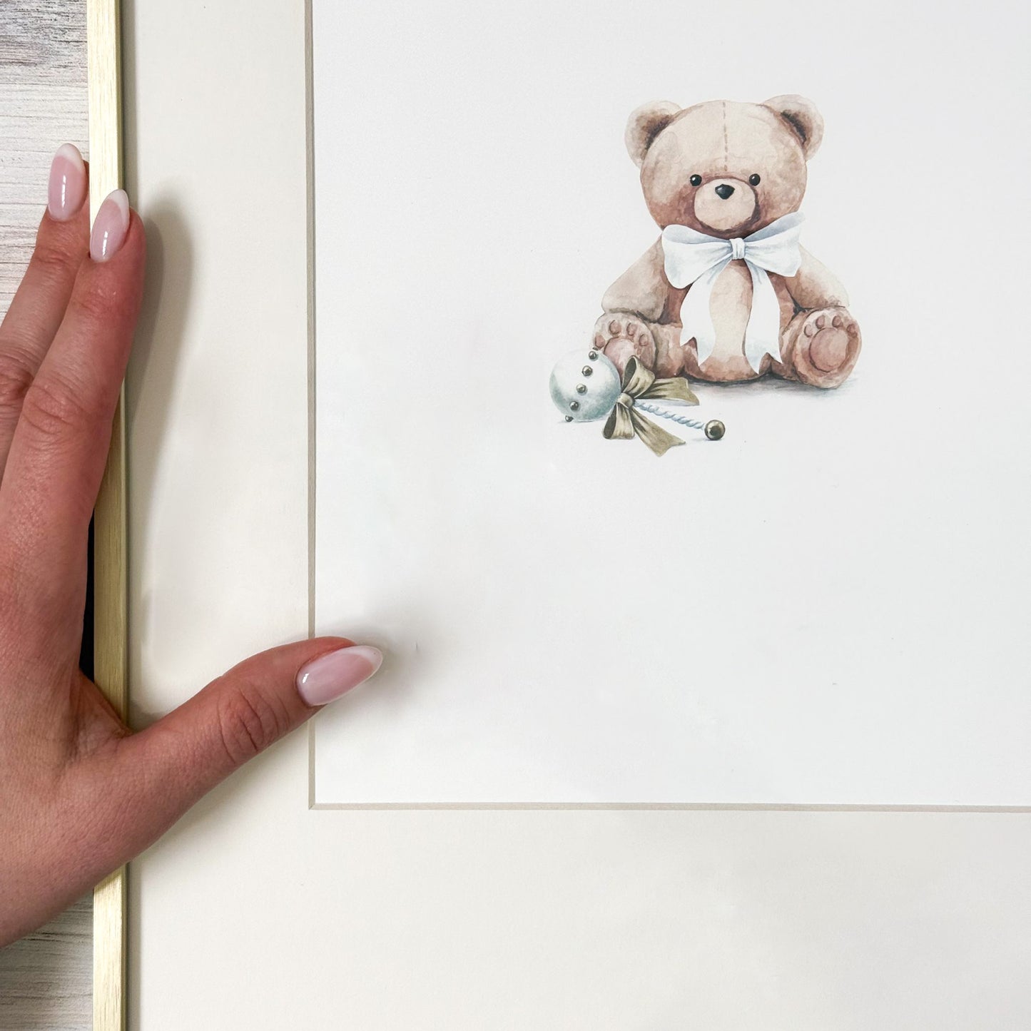 Hand holding a white frame with a teddy bear and bird illustration on a light wooden surface.