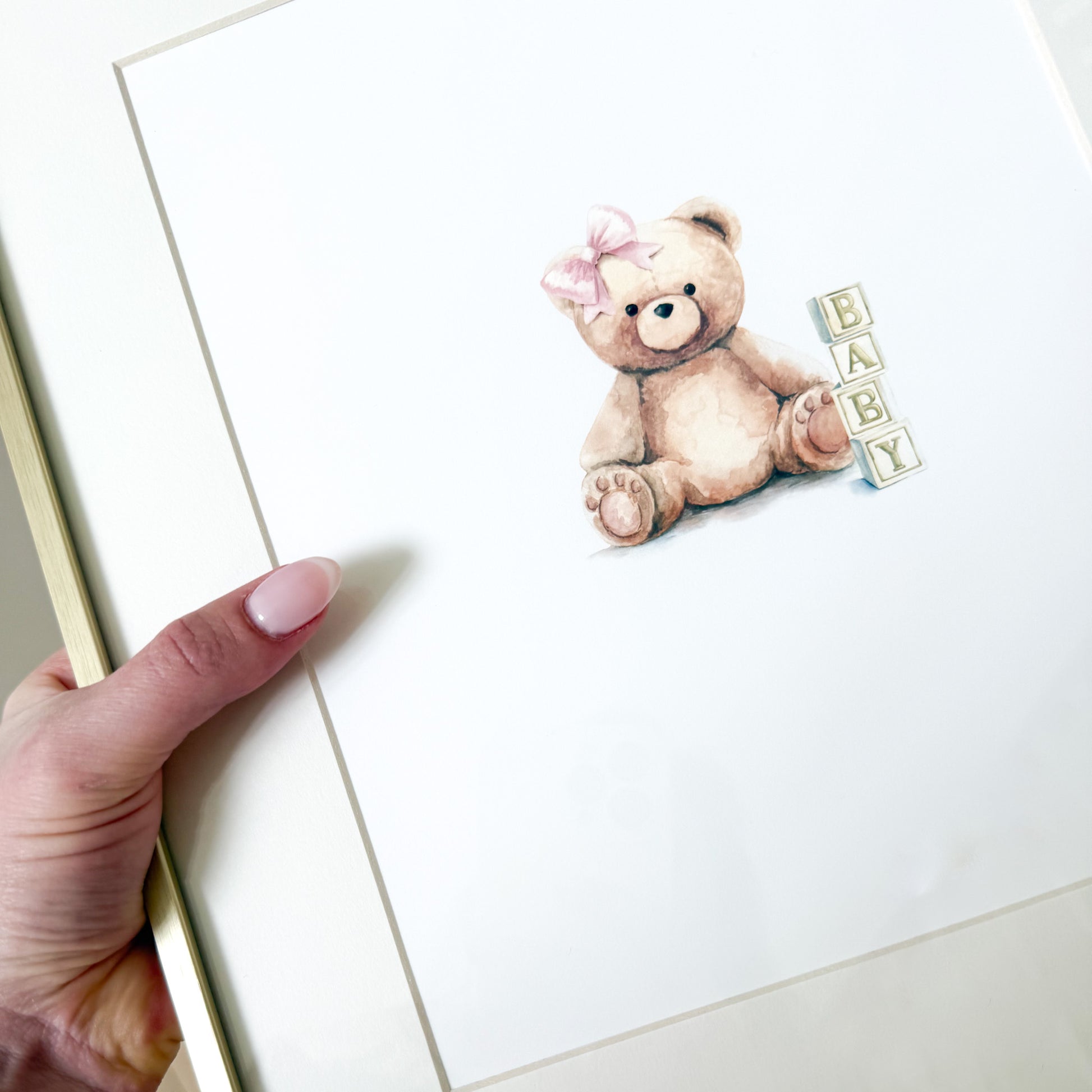 Hand holding a gold frame with a watercolor teddy bear with a pink bow and blocks that read BABY