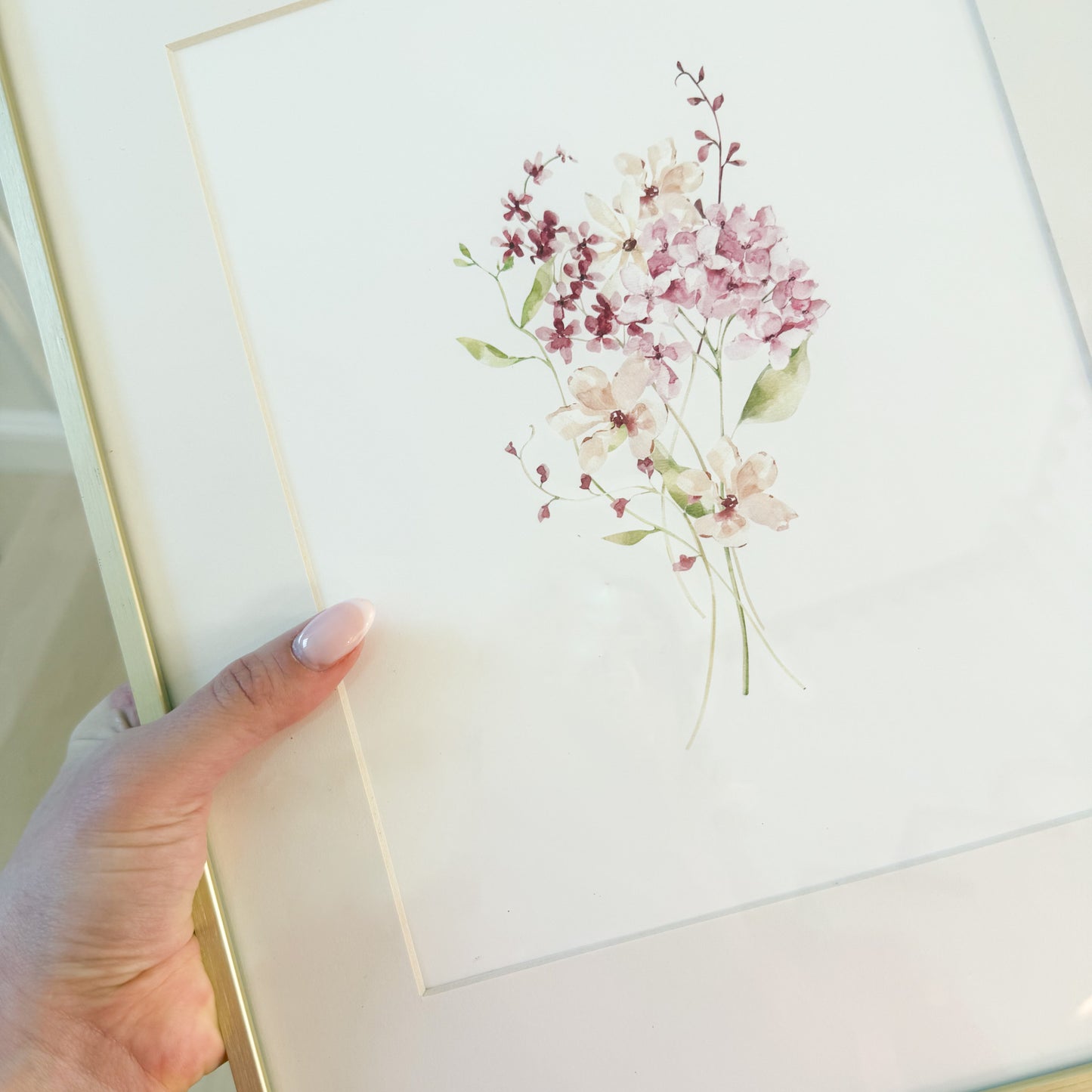 hand holding a gold frame with a picture of a watercolor wildflowers bouquet with pink and burgundy flowers.