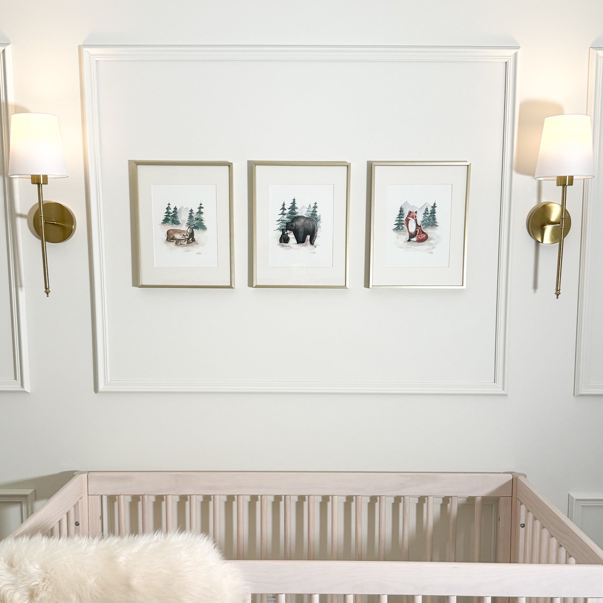 Watercolor art prints of deer and fawn, black bear and cub, and fox and baby fox displayed above a crib in gold frames.
