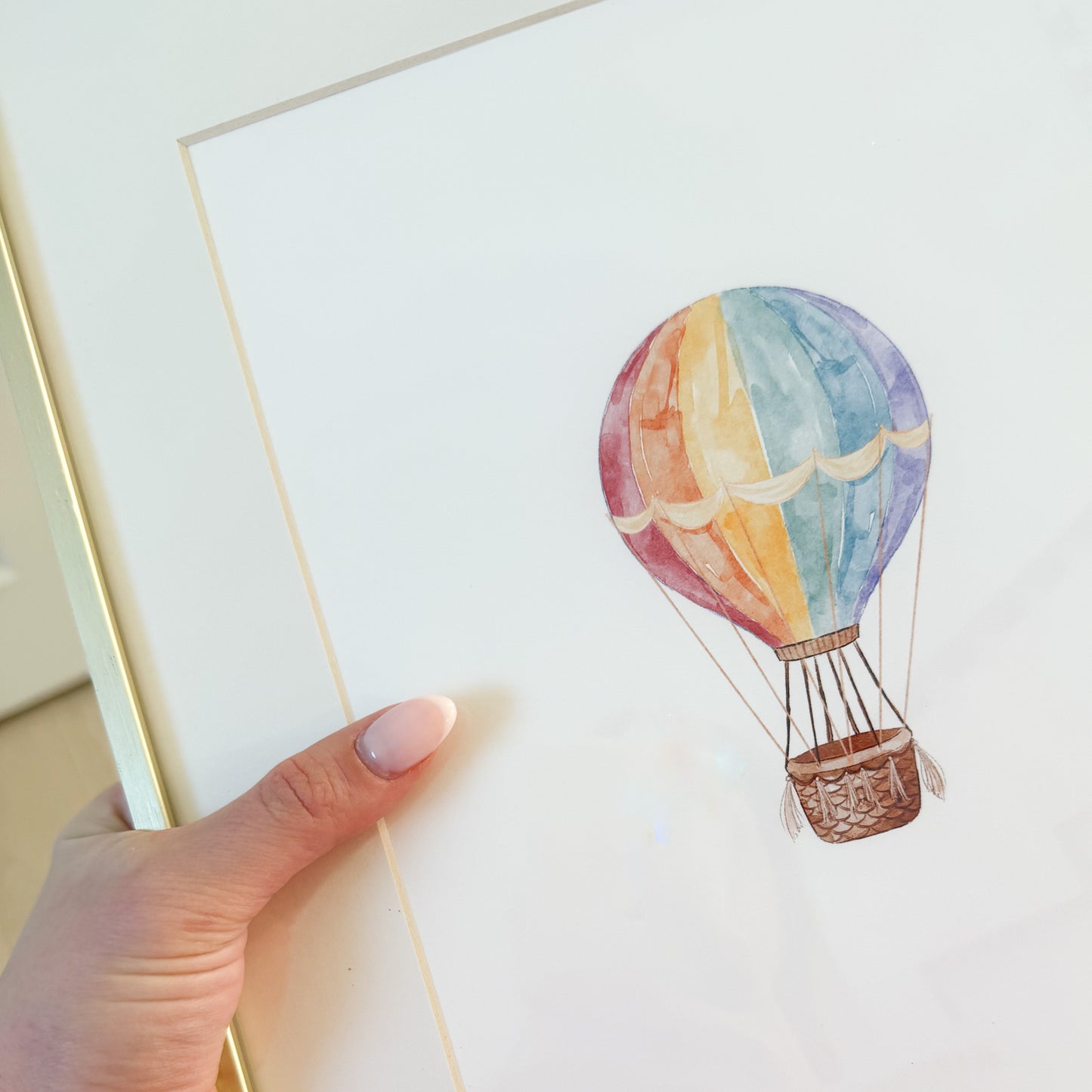 Rainbow hot air balloon done in watercolor against a white background in a gold frame.