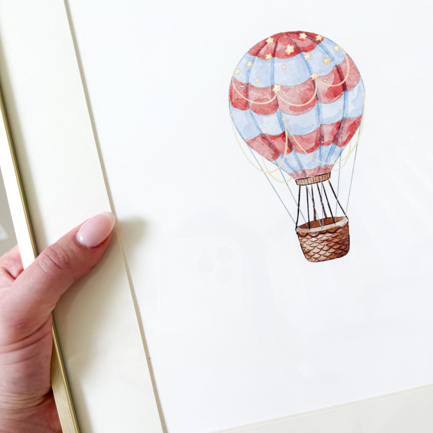 Hand holding a gold frame with a red and blue watercolor hot air balloon on a white background