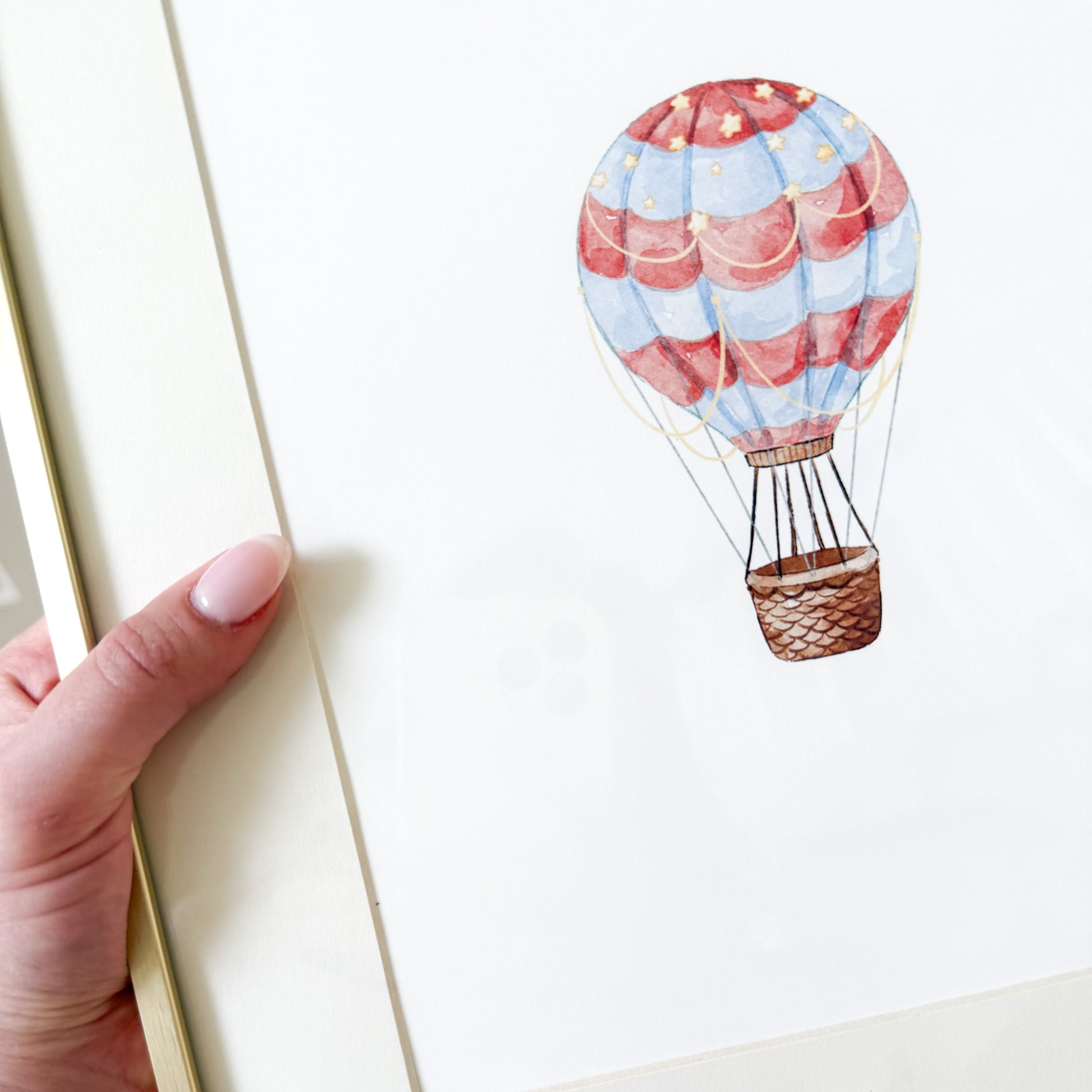 Hand holding a gold frame with a red and blue watercolor hot air balloon on a white background
