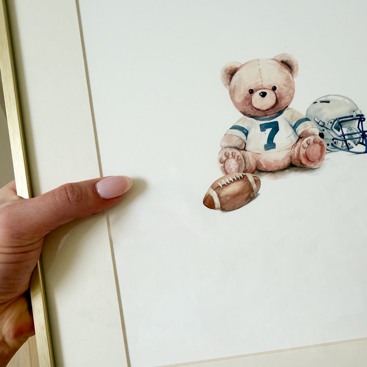 Watercolor teddy bear in vintage football gear with a helmet and football against a white background within a gold frame.