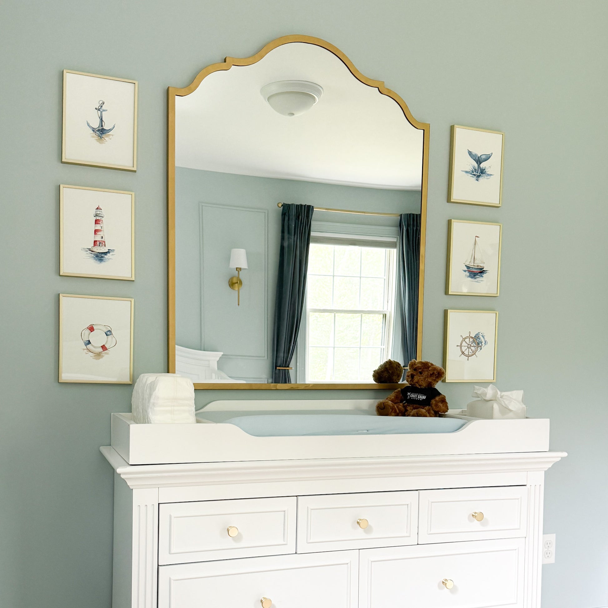 A large arched gold-framed mirror mounted on a pale blue wall above a white dresser, with framed nautical prints on both sides and a changing tray and teddy bear on the mirror surface.