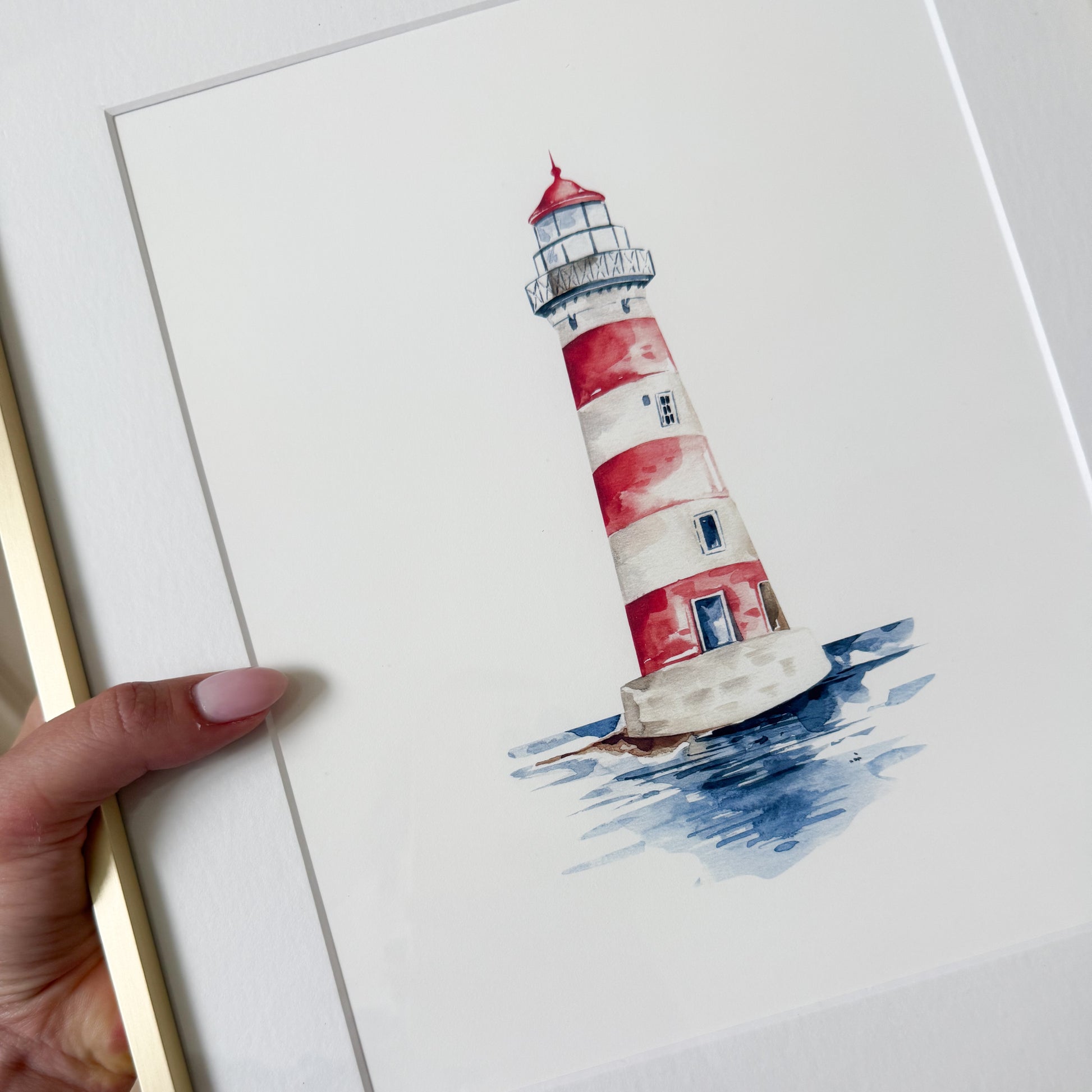 A framed watercolor art print of a red and white striped lighthouse standing in blue water, held by a hand. The print is mounted on a white mat board with a gold-toned frame visible on the left.