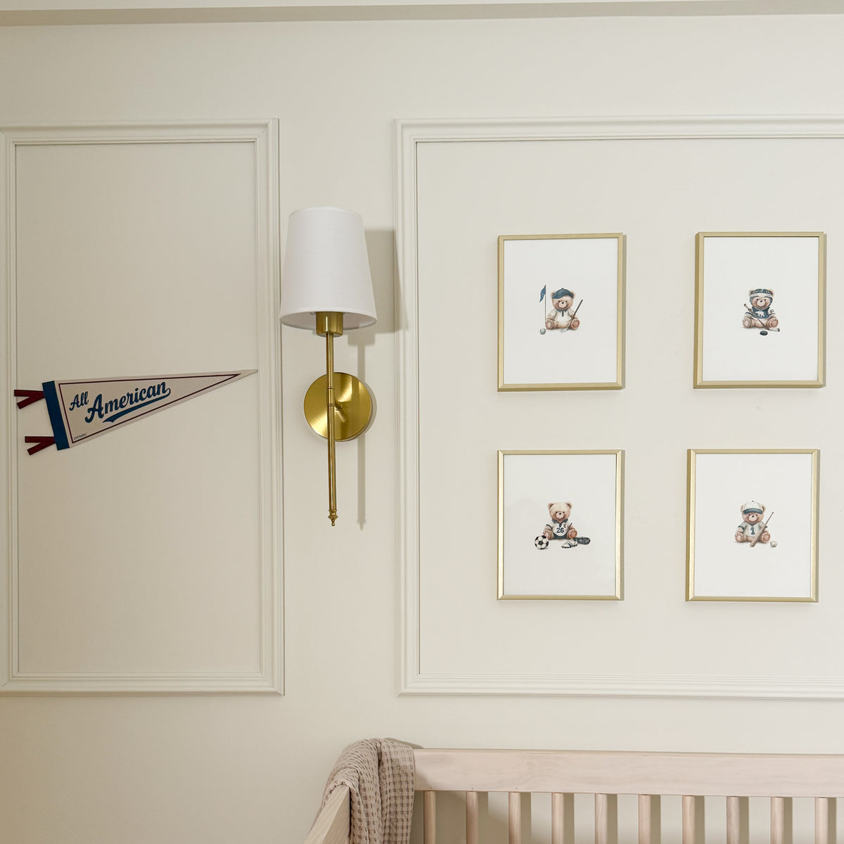A styled nursery wall featuring a gold and white wall sconce, a white panelled border, a small 'All American' pennant, and four framed teddy bear illustrations above a crib.
