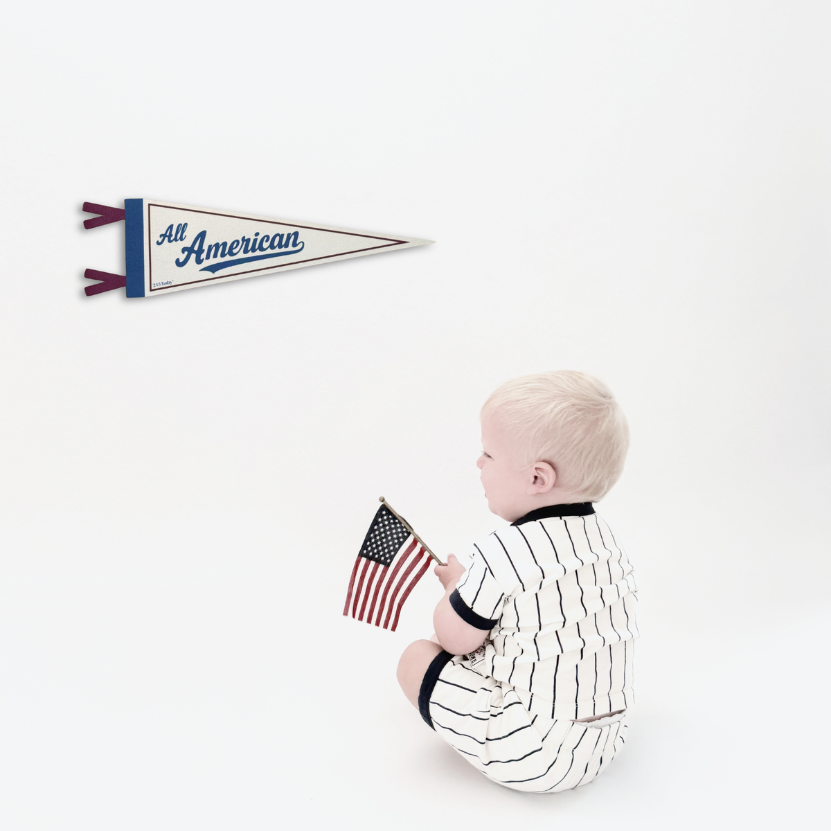 A baby in a navy and white pinstripe outfit sits on a white background holding a small American flag, with a matching 'All American' pennant displayed on the wall above.
