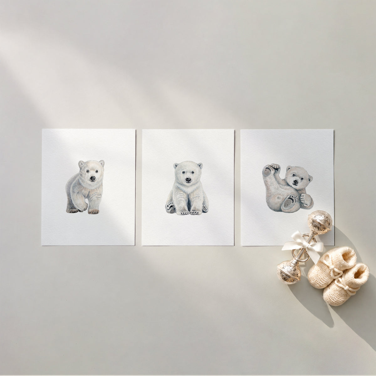 Set of 3 unframed watercolor polar bear cub nursery art prints featuring a walking cub, a seated cub, and a playful cub rolling on its back, flat laid in a row with cream knit baby booties and a silver rattle.