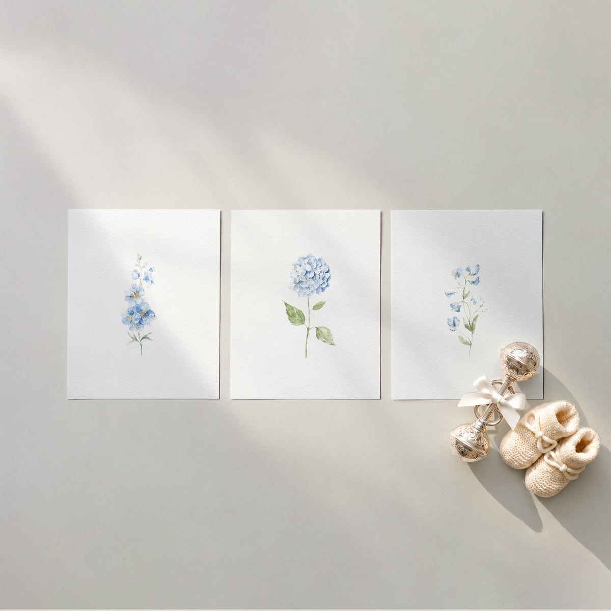 Set of 3 unframed watercolor light blue botanical nursery art prints featuring delphinium, blue hydrangea, and sweet pea stems, flat laid in a row with cream knit baby booties and a silver rattle.