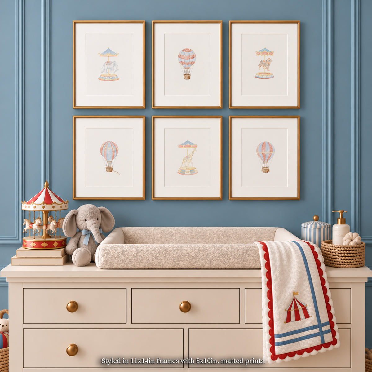 A set of six gold-framed carousel and hot air balloon watercolor nursery wall art prints displayed in a 2x3 grid on a dusty blue paneled wall above a cream changing dresser, styled with a plush elephant toy, toy carousel, and a red and blue trimmed blanket.