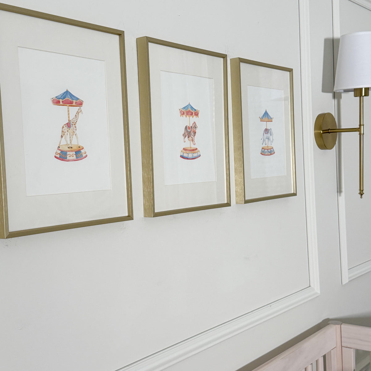 Three framed art prints featuring whimsical carousel illustrations in gold frames, displayed on a white wall next to a brass wall sconce.