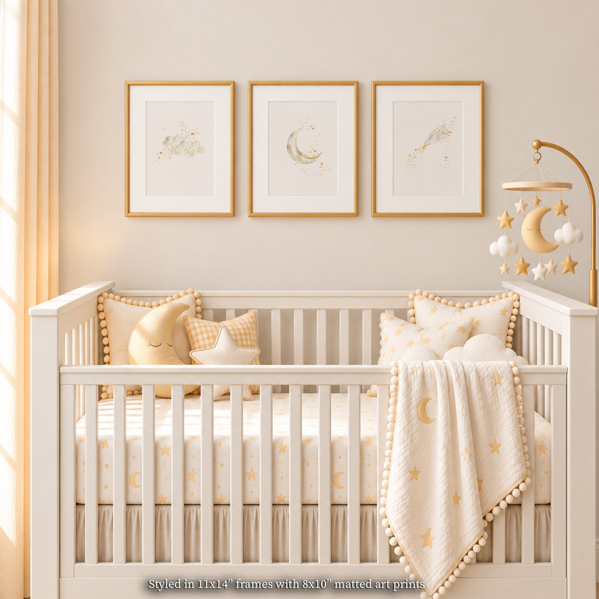 Cozy neutral and gold nursery mockup featuring a row of 3 matted gold-framed watercolor celestial prints (clouds, crescent moon, shooting star) centered above a white wood crib. Styled with soft yellow and cream star-patterned bedding, celestial pillows, a pom-pom trimmed blanket, and a gold baby mobile, with natural linen drapery.
