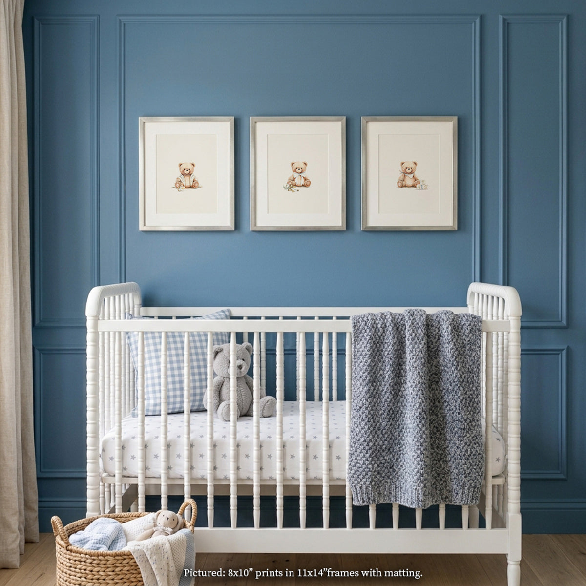 Close-up boys nursery gallery wall mockup featuring a row of 3 matted silver-framed watercolor teddy bear prints centered above a white spindle crib. Styled against a rich slate blue panelled wall with picture frame moulding, the crib features star-patterned sheets, a blue gingham pillow, a textured heather-blue chunky knit blanket, and a grey plush teddy bear, with a basket of baby items on the floor.