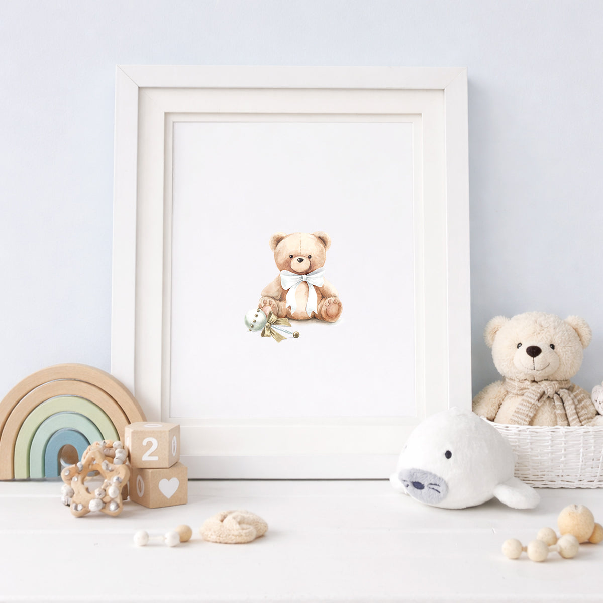 Framed watercolor art print of a classic teddy bear wearing a ribbon bow, styled on a light blue nursery shelf with a wooden rainbow stacker and plush bear.