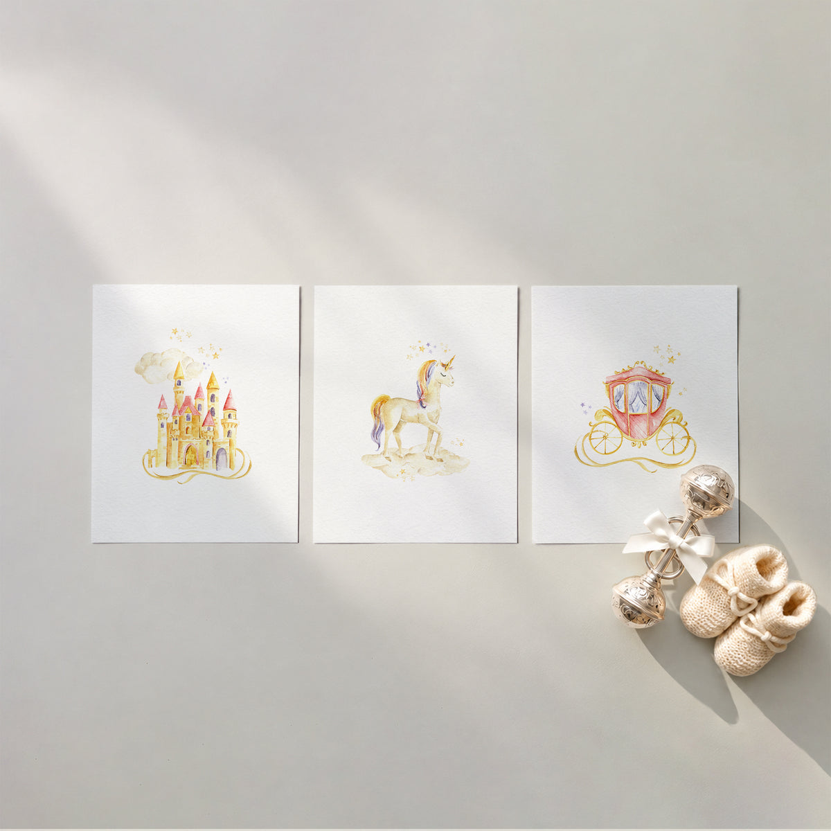 Set of 3 unframed storybook fairytale watercolor nursery art prints featuring a pink and gold fairytale castle, a magical unicorn, and a royal carriage, flat laid in a row with cream knit baby booties and a silver rattle.
