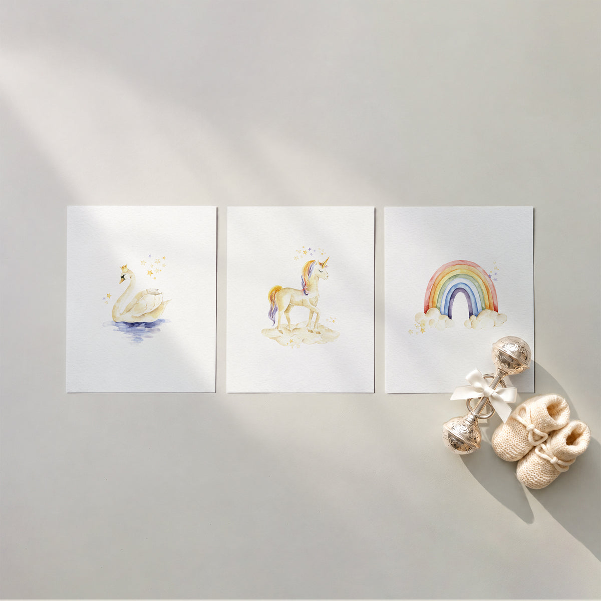 Set of 3 unframed storybook fairytale watercolor nursery art prints featuring a crowned swan, a magical unicorn, and a pastel rainbow over clouds, flat laid in a row with cream knit baby booties and a silver rattle.