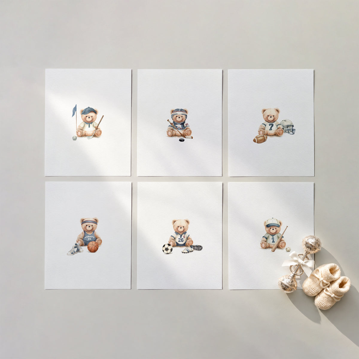 Flat lay composition of 6 sports teddy bear watercolor art prints on a warm neutral surface with knitted baby booties and a silver rattle.