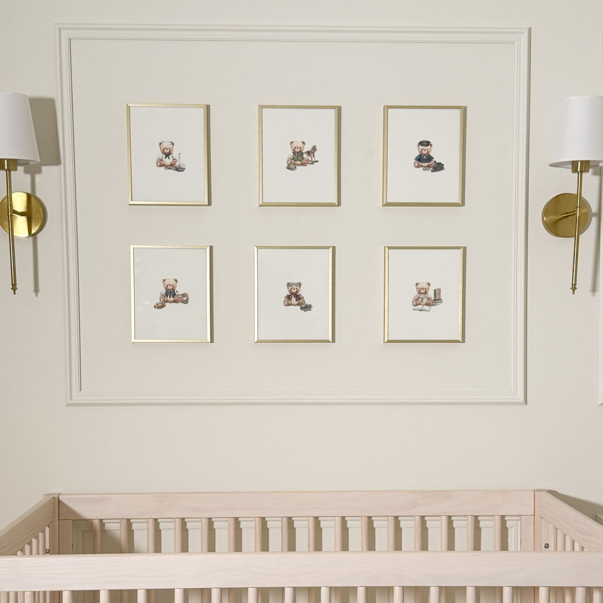A nursery wall featuring six small framed illustrations of teddy bears in a 2x3 grid, mounted above a light wood crib. The wall is cream-colored with a decorative molding border, flanked by two gold wall sconces.