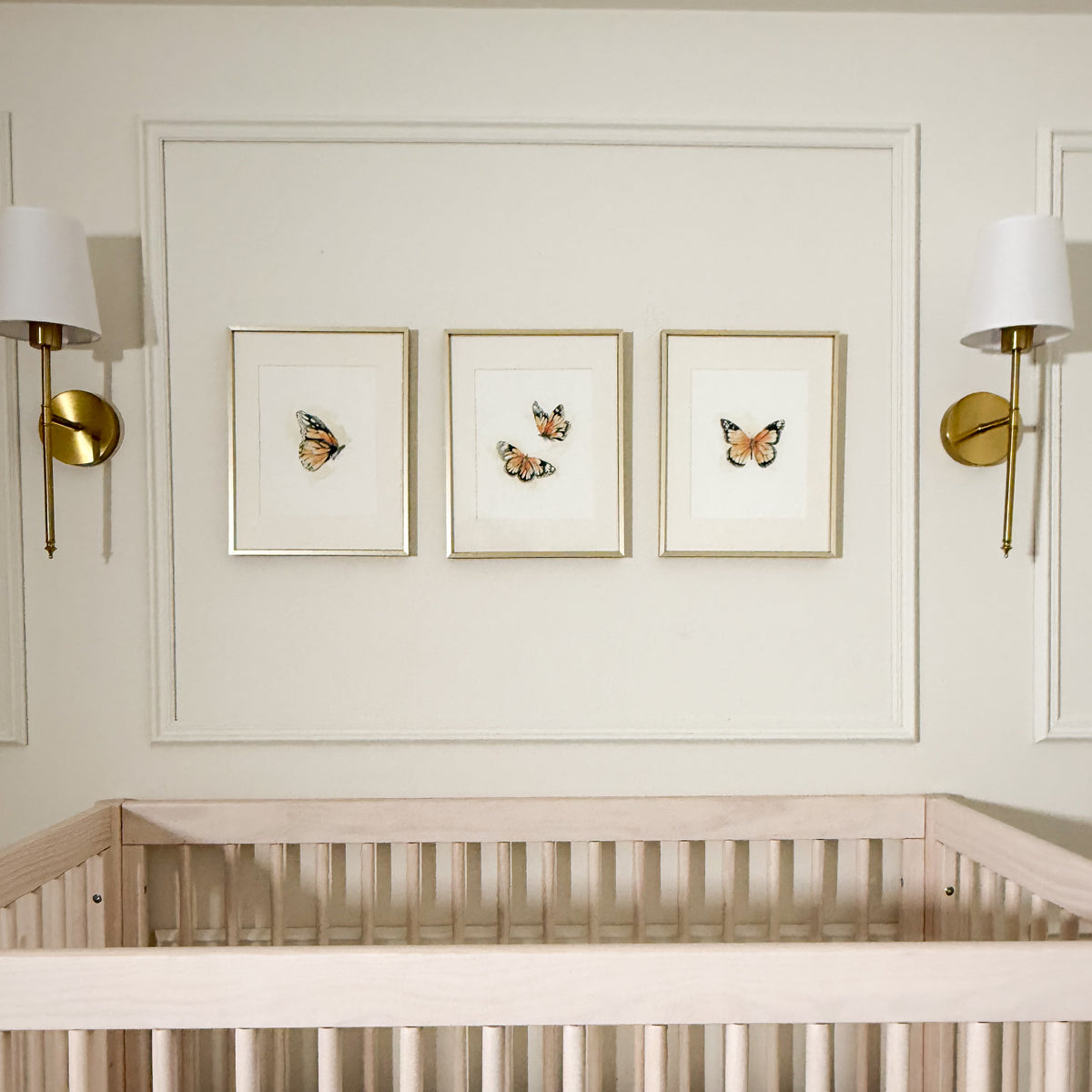 Three framed butterfly art prints with gold frames hang on a cream wall above a light wood crib, flanked by two gold wall sconces.