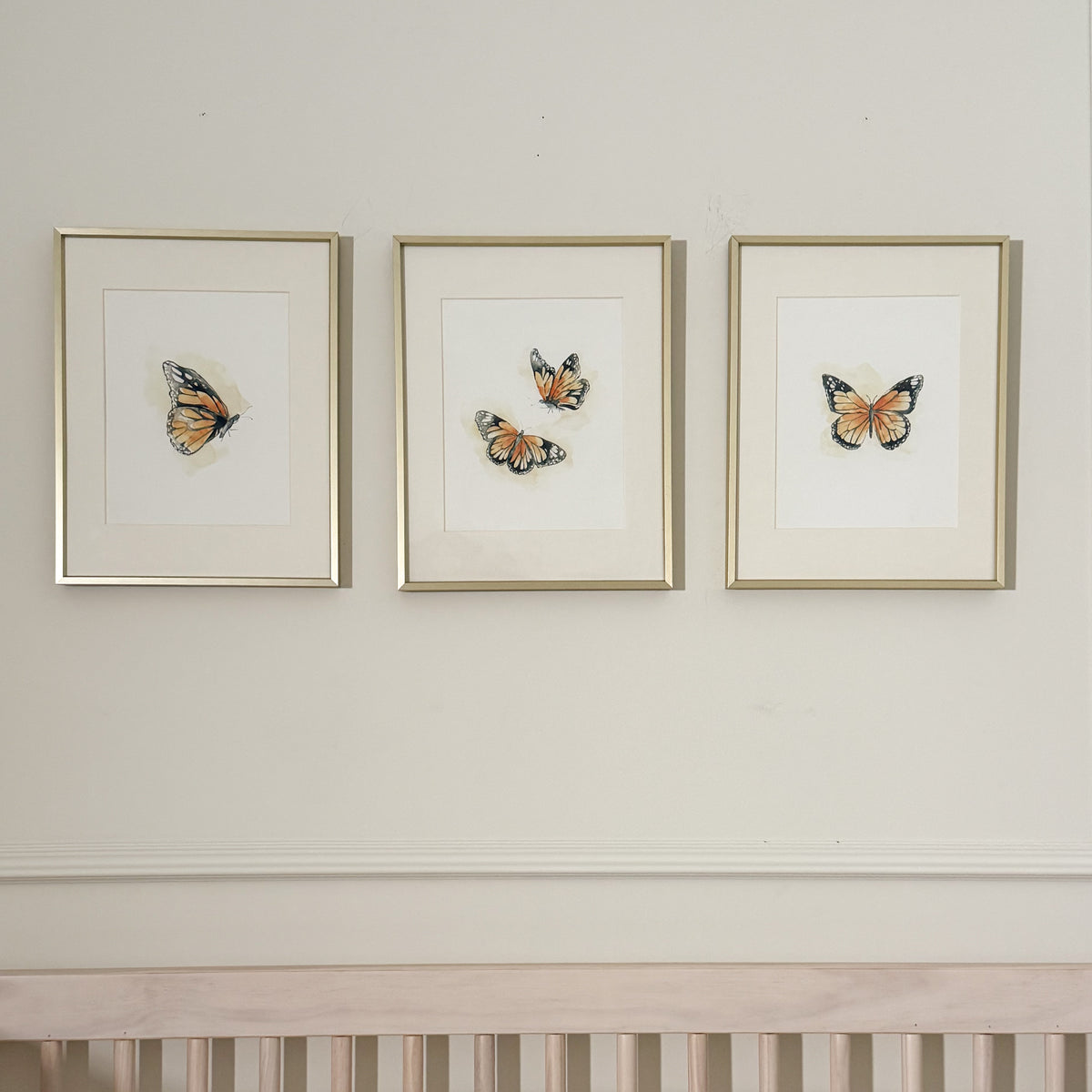 Three framed butterfly art prints with gold frames hanging on a cream wall above a light wood crib.