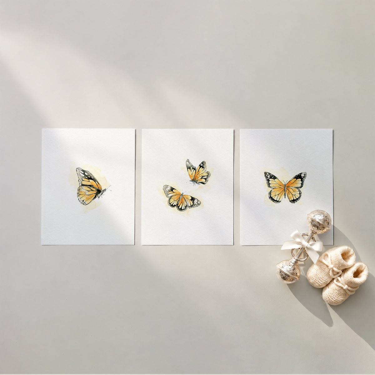 Set of 3 unframed watercolor monarch butterfly nursery art prints featuring yellow and orange gold butterflies in side profile, a pair, and open-wing view, flat laid in a row with cream knit baby booties and a silver rattle.