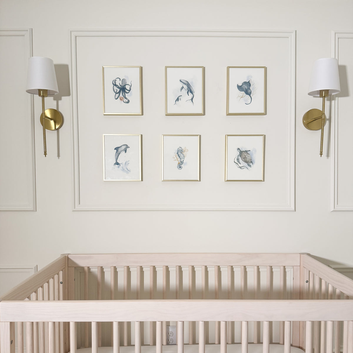 A styled nursery wall featuring a gallery wall of six framed watercolor prints of sea life, flanked by two gold and white wall sconces above a light wood crib.