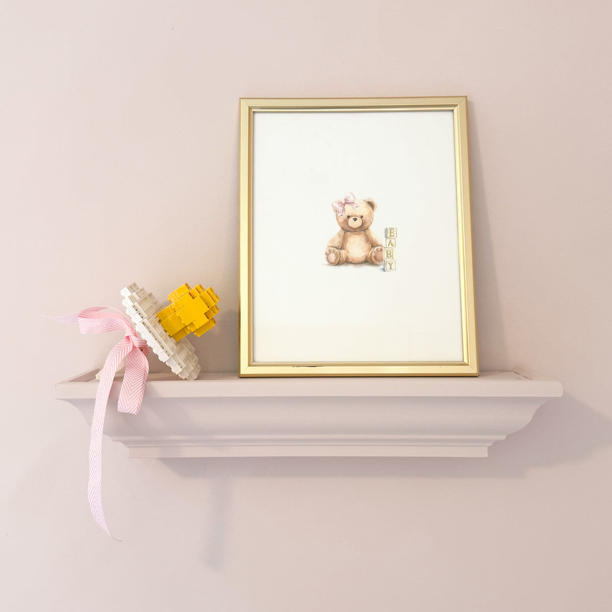 A framed art print featuring a small watercolor teddy bear with a pink bow, displayed on a white shelf next to a yellow and white decorative object against a soft pink wall.