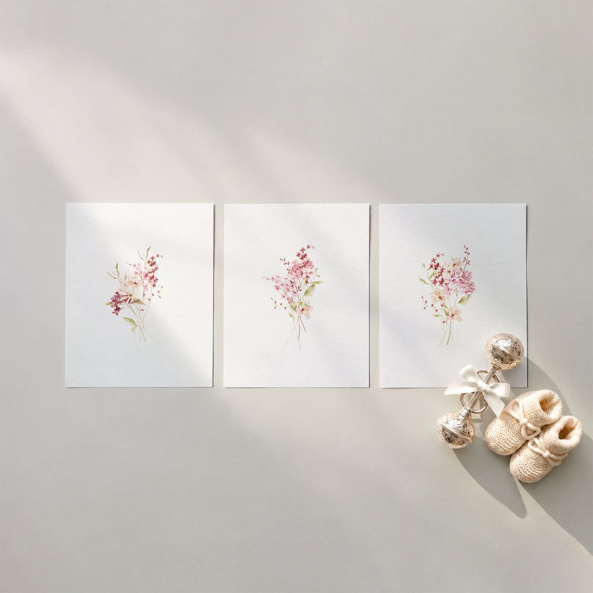 Flat lay mockup of 3 pink wildflower watercolor nursery art prints arranged on a warm neutral surface with baby booties and a silver rattle.