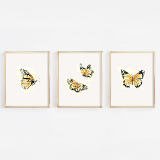 Set of three 8x10 gold-framed nursery prints featuring watercolor monarch butterflies with a soft orange glow, perfect for a gender-neutral or nature-themed baby room.