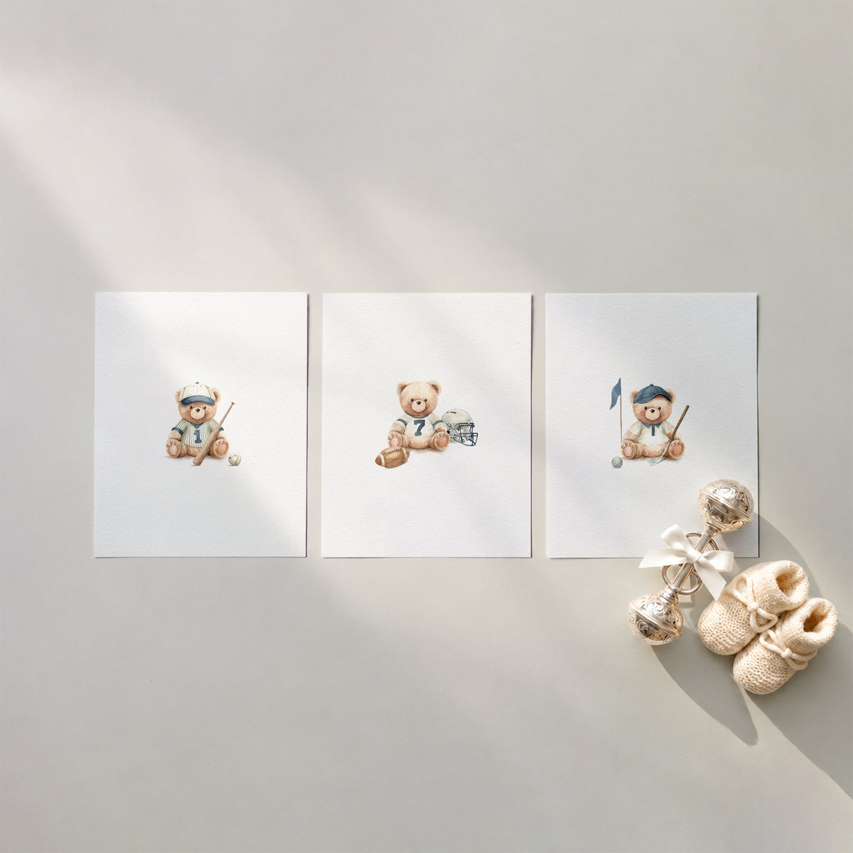 Flat lay mockup of 3 sports teddy bear watercolor nursery art prints featuring baseball, football, and golf designs arranged on a warm neutral surface with baby booties and a silver rattle.
