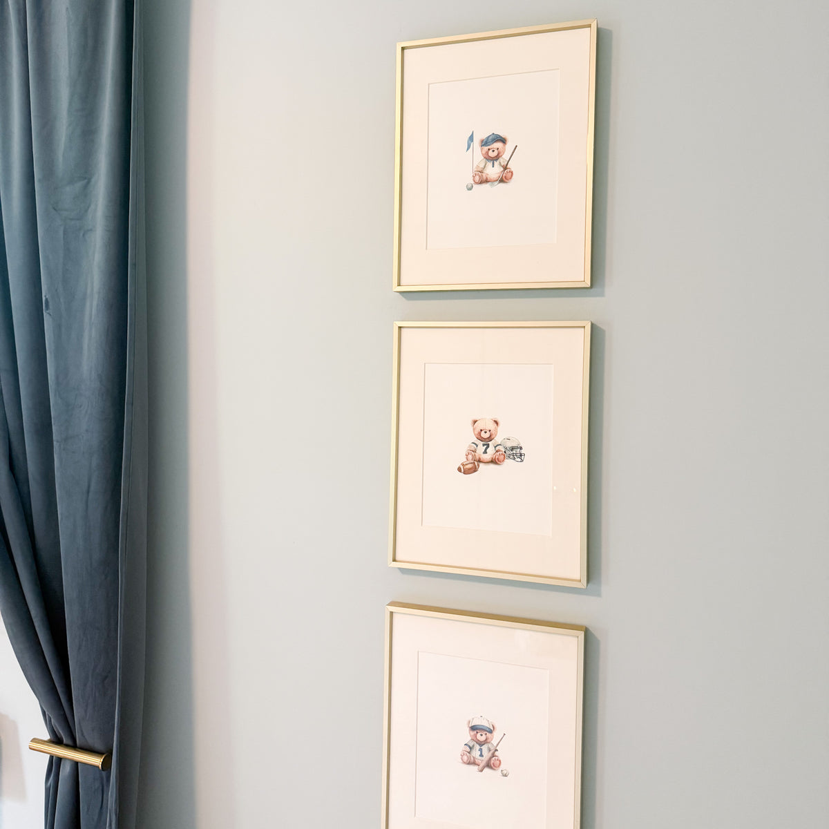 Three vertically arranged framed art prints with cream mats and thin gold frames hang on a light gray wall next to a blue curtain. Each print features a small  teddy bear illustration in sports outfits for baseball, football, golf.