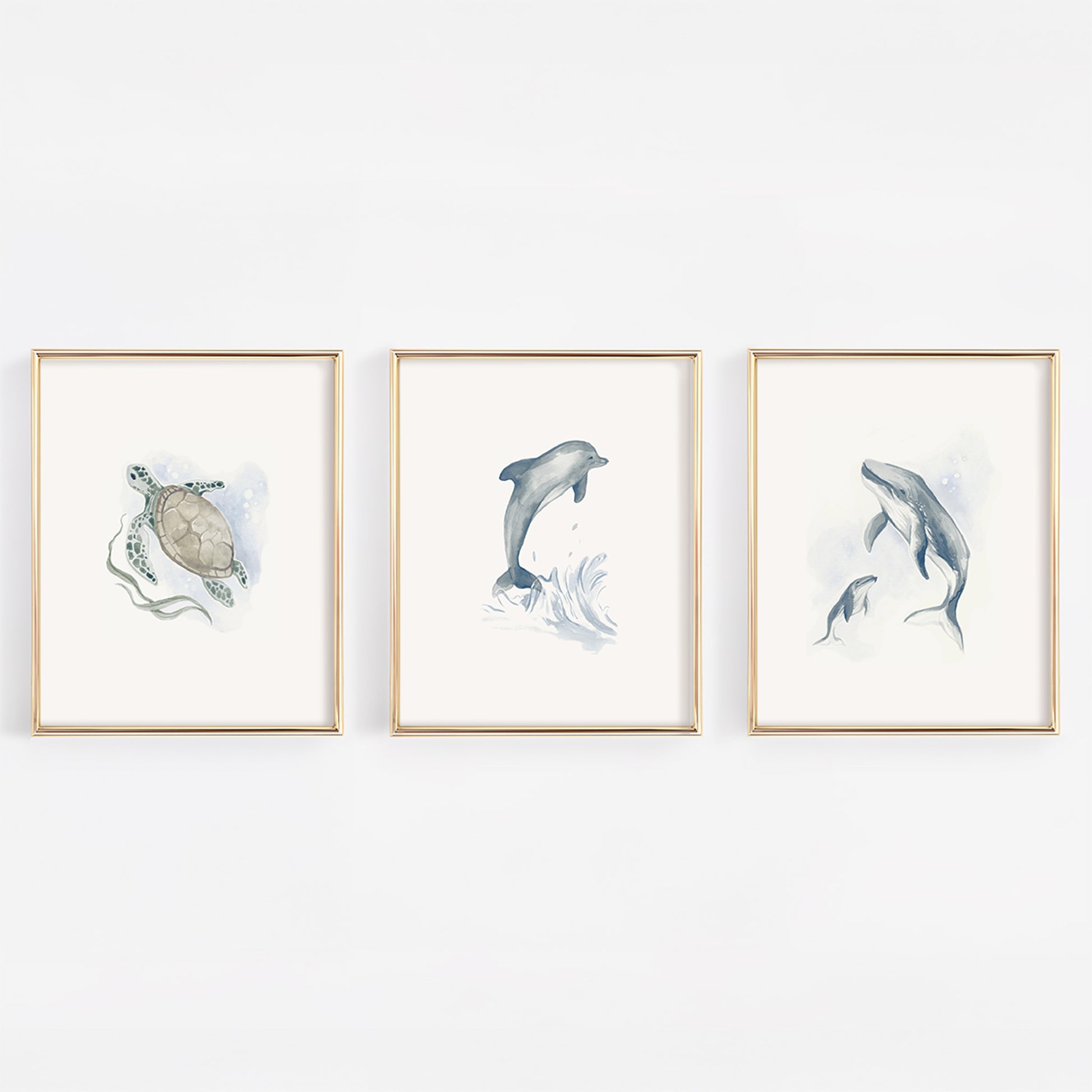 Set of three gold-framed nursery prints featuring watercolor marine life: a gentle sea turtle, a mother and baby humpback whale, and a playful blue dolphin on a clean white background.