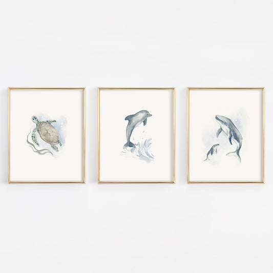 Set of three gold-framed nursery prints featuring watercolor marine life: a gentle sea turtle, a mother and baby humpback whale, and a playful blue dolphin on a clean white background.