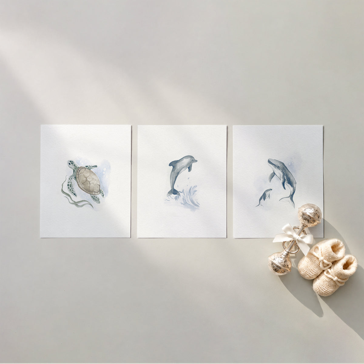 Set of 3 unframed watercolor under the sea ocean nursery art prints featuring a sea turtle, a leaping dolphin, and a mother and baby humpback whale, flat laid in a row with cream knit baby booties and a silver rattle.