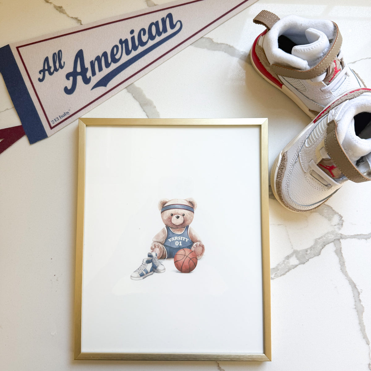 A framed art print featuring a teddy bear basketball illustration, styled on a white marble surface with a gold frame, white sneakers, and a 'All American' pennant.