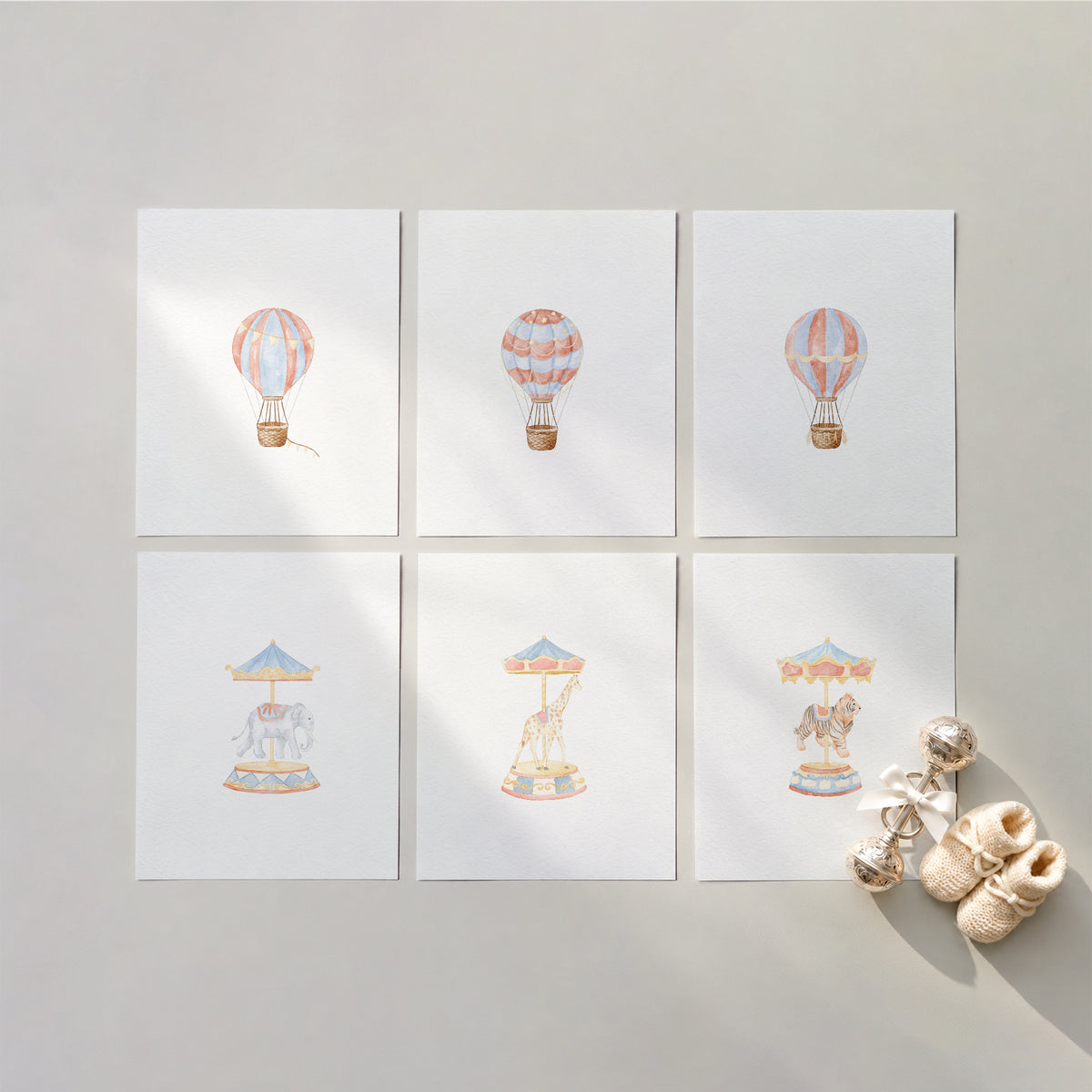 Flat-lay composition of six unframed watercolor nursery art prints on textured cotton fine art paper arranged in a 2x3 grid on a neutral background, featuring vintage hot air balloons and carousel animals with a silver baby rattle and knit booties.