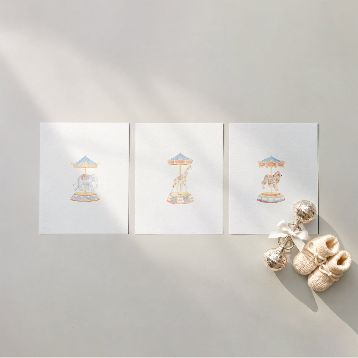 Set of 3 unframed watercolor carousel animal nursery art prints featuring a carousel elephant, giraffe, and tiger under decorative tops, flat laid in a row with cream knit baby booties and a silver rattle.