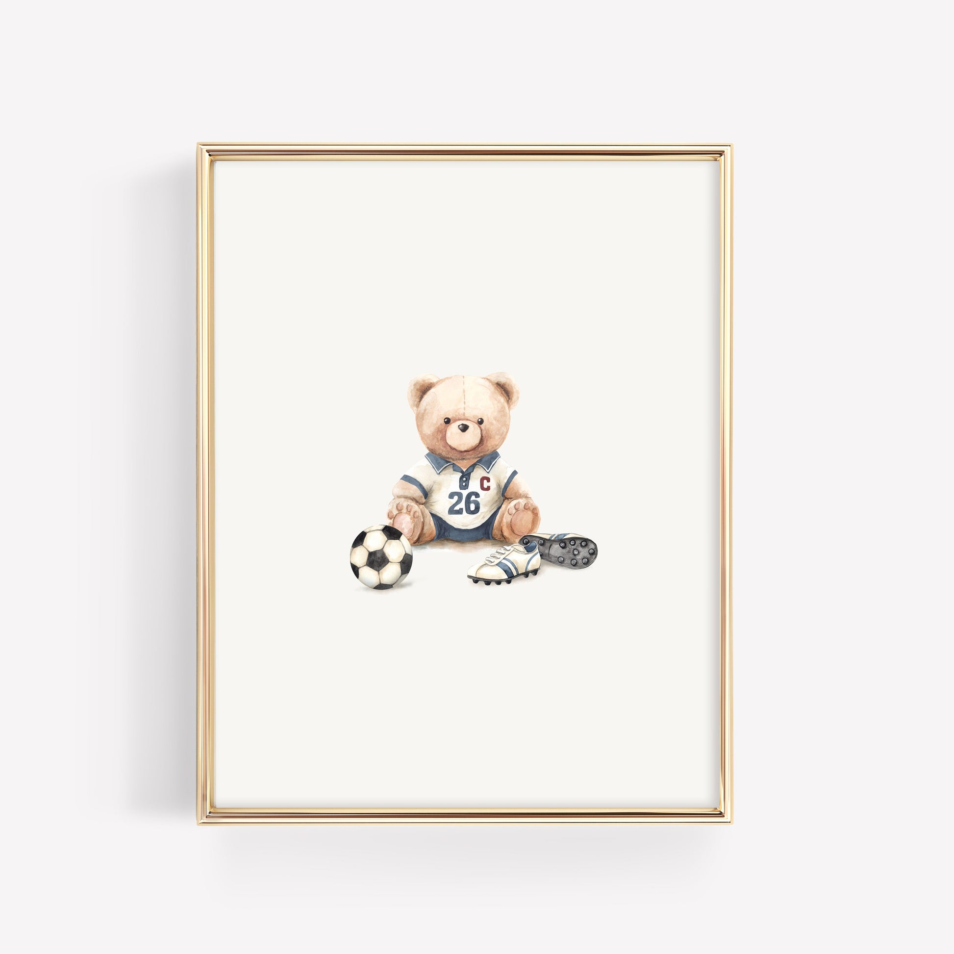 A framed art print featuring a teddy bear wearing a blue soccer jersey with the number 26, sitting next to a soccer ball, a cleat, and a small ball, displayed in a thin gold frame against a light gray background.