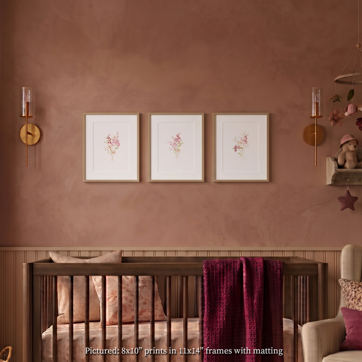 Set of 3 framed wildflower watercolor art prints in light wood frames with white mats, hanging on a warm taupe nursery wall above a dark wood crib with a burgundy blanket.