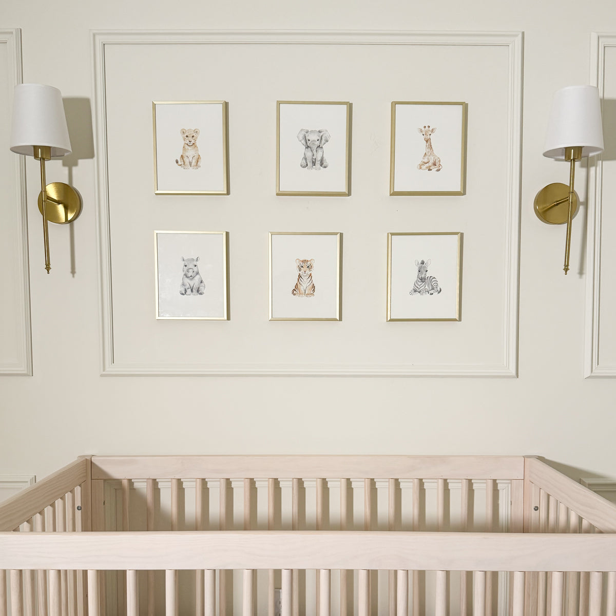 A nursery wall featuring six framed animal illustrations arranged in two columns of three above a light wood crib. The wall is warm off-white with two gold and white wall sconces on either side of the framed art.