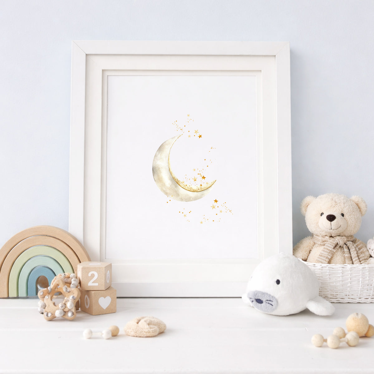 Framed watercolor art print of a soft crescent moon and sparkling stars, styled on a neutral nursery shelf with a wooden rainbow toy and teddy bear.