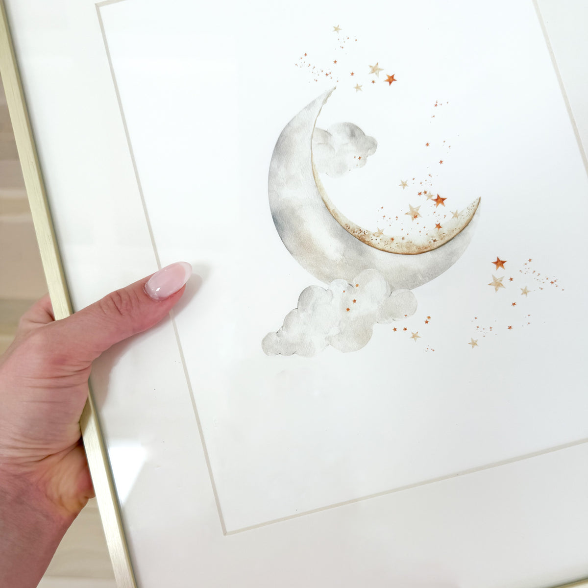 A watercolor painting of a silver moon and gold stars against a white background in a gold frame.