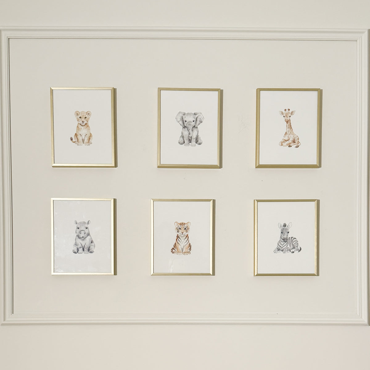 A gallery wall display featuring six small framed animal illustrations arranged in two rows of three. The prints show a lion cub, baby elephant, baby giraffe, baby tiger, baby zebra, and baby rhino in soft watercolor styles, each in a thin gold frame on white backgrounds.