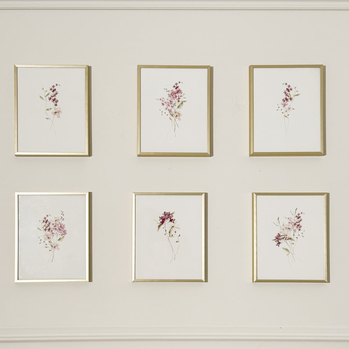 Six framed botanical watercolor prints arranged in two rows of three on a light beige wall. Each print features delicate pink, purple, and green floral illustrations on a white background, set in thin gold frames.