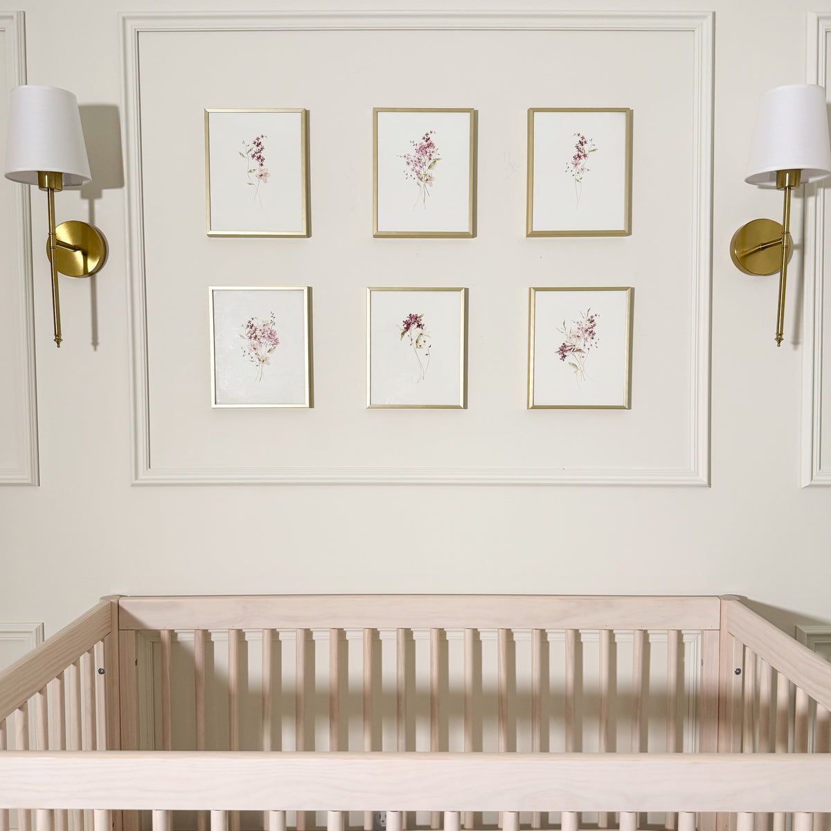 A styled nursery interior showing a light wood crib in the foreground, a cream wall with decorative molding, six small framed floral prints, and two gold wall sconces.